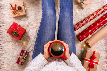 Young woman drinking coffee and sitting on the furry rug. Close-up of female legs in warm socks with a deer print, wrapping paper rolls, top view. Christmas preparation concept. Background, copy space