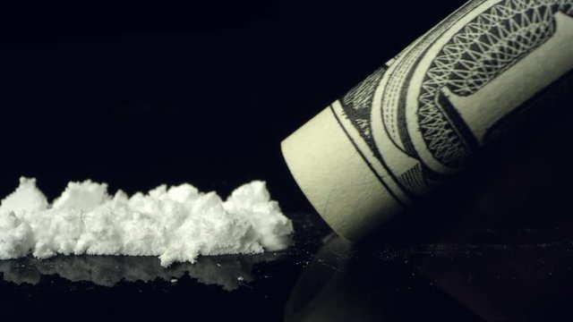 View of white powder drug being snorted up rolled up dollar bill on black reflective table top.
