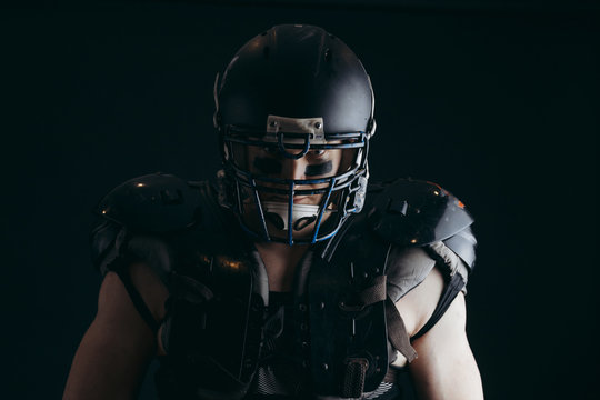 Head and shoulder portrait of muscular American football player wearing black helmet and protective armour over naked torso, looking at camera over black background