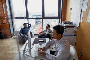Smiling asian businessman in formal clothes typing on laptop in office, while his coworkers chatting near the big panoramic window