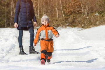 Little boy in overalls runs away from his mother. Winter snowy day in the coniferous forest