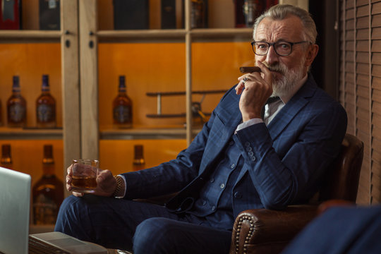 Senior cute business man with cigar and whisky. Gray hair and beard wearing blue tailored suit and tie, relaxing at fashionable restaurant lounge