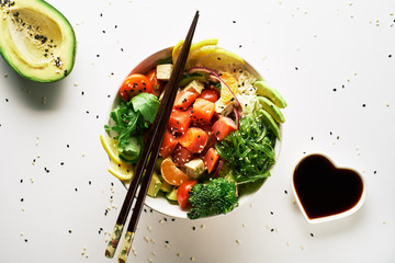 poke bowl with salmon, avocado, cucumber, arugula, broccoli, rice, carrot and sweet onions with chuka salad, with chopsticks, avocado and soy sauce isolated over white background. top view