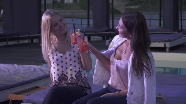 Two delicious girls drinking cocktails in a club. Two smiling girlfriends clink glasses with cocktails and chat sitting in a summer club. Two cute girls sitting in a night cafe spend time together.