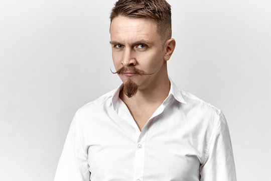 Isolated picture of handsome successful young male entrepreneur with handlebar mustache and goatee beard posing in studio wearing white formal shirt, looking at camera with confident smile