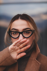 Attractive woman walking near business center windows. Photo of  blonde female model looks with cover mouth. Concept of warm and comfort. Girl wear glasses, coat, sweater with neck. 