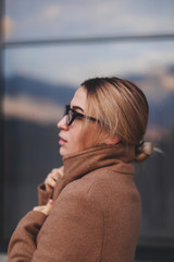 Attractive beauty woman walking near business center while wearing beige coat and glasses. Photo blonde girl which turn around. The window is a reflection of the sky.