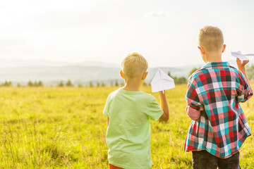 two blonde boys are playing with paper airplanes in a meadow, having fun