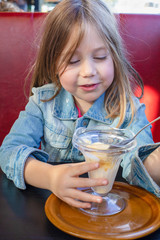 Portrait of five years old blonde little child with blue denim jacket, sitting in red sofa and black table in a restaurant, eating vanilla ice cream with spoon from crystal cup. vertical shot