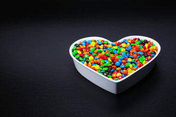 White shaped ceramic bowl filled with small colorful round coated candies on a black background with text space. Concept:  loving sweets and candy.  