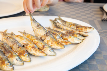 hand of little girl catching a sardine cooked from the tail in a big white dish ready to eat on table of restaurant