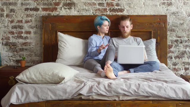 Medium shot of young woman with blue hair and hipster man with beard sitting on bed in loft apartment and talking while looking at laptop