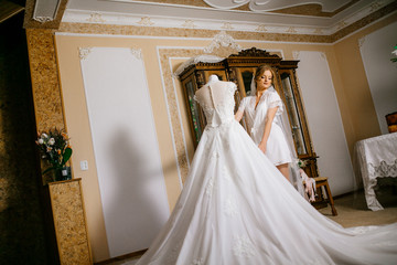 Obraz na płótnie Canvas Attractive bride in white robe, long veil standing near the wedding dress on the mannequin on wedding morning. Preparing for the ceremony
