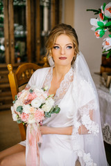 Gorgeous beautiful bride in silk robe and veil holding modern bouquet posing in light. Happy stylish Bride with perfect hairstyle posing near window in the morning.