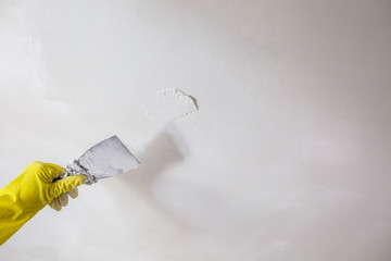worker's hand in yellow gloves holding putty knife patching a hole with spatula with plaster or...