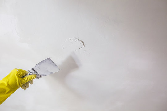 worker's hand in yellow gloves holding putty knife patching a hole with spatula with plaster or putty in white wall. Renovation and repair process, remodeling interior of room at apartment building