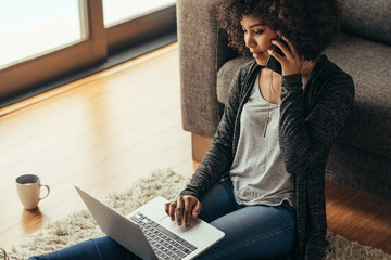 African woman at home using laptop and talking on phone