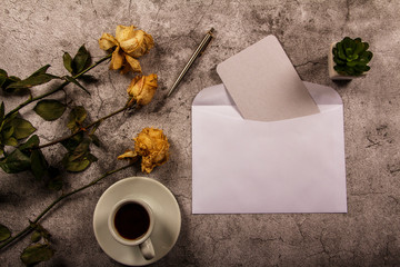 Mock up template with wilted flowers of white roses with a clean envelope, notepad and cup of coffee on a gray concrete background. View from above. Flat lay. Template layout for designer.