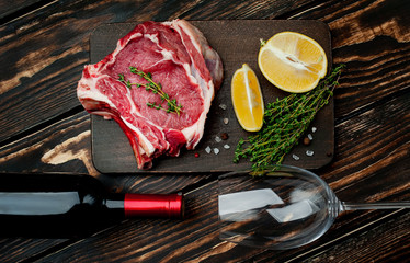 Raw steak with a bottle of wine and a glass, lemon, red pepper, garlic and seasonings on a dark rustic background
