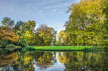 Fototapeta na wymiar Trees in various colors, a blue sky, reflection in a mirror-like pond and a green lawn in the Park in Rotterdma on a sunny day in autumn