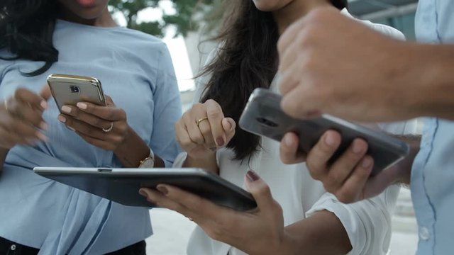 Three people standing outside with electronic devices, gesticulating actively. Three colleagues holding tablet PC and two smartphones, communicating. Close up footage. Technology concept.