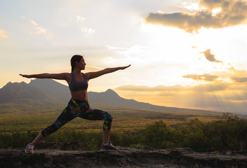 Silhouette of young woman practicing yoga or pilates at sunset or sunrise in beautiful mountain location.