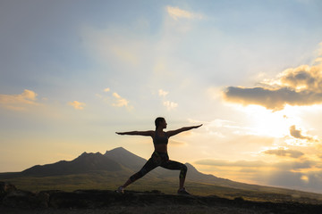 Silhouette of young woman practicing yoga or pilates at sunset or sunrise in beautiful mountain location.
