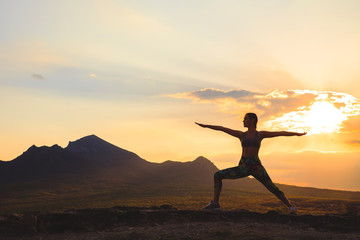 Silhouette of young woman practicing yoga or pilates at sunset or sunrise in beautiful mountain location.