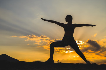 Silhouette of young woman practicing yoga or pilates at sunset or sunrise in beautiful mountain location.