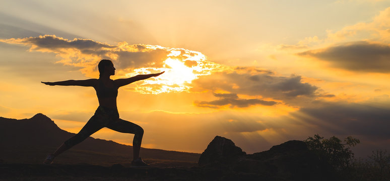 Silhouette of young woman practicing yoga or pilates at sunset or sunrise in beautiful mountain location.