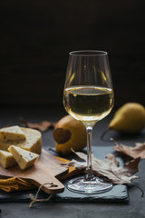 A glass of white wine served with cheese in a cutting board on dark background