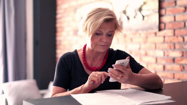Senior businesswoman comparing documents with smartphone by table at home

