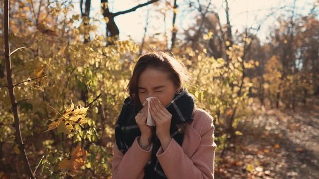 Pretty girl is caught cold and sneezing during walk in forest in autumn day. She is wrapped in warm scarf, holding hanky and wiping her nose