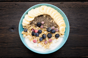 Healthy fitness breakfast: oatmeal with bananas, blueberries and cranberries, chia seeds and yogurt on dark wooden background with texture