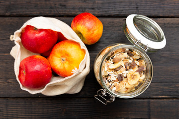 Homemade granola in glass jar and apples in reusable fabric bag on dark wooden table with texture. Healthy breakfast concept and zero waste concept