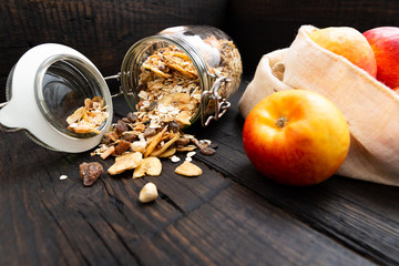 Homemade granola in glass jar and apples in reusable fabric bag on dark wooden table with texture. Healthy breakfast concept and zero waste concept