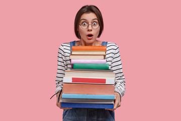 Astounded young European woman with dark hair, dressed in striped clothes, carries many books, being stupefied to learn much information for exam, isolated over pink background. Studying concept.