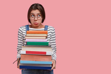 Caucasian displeased young college student holds pile of books, prepares for examination session, purses lips in discontent, dressed in striped sweater and overalls, isolated over pink background