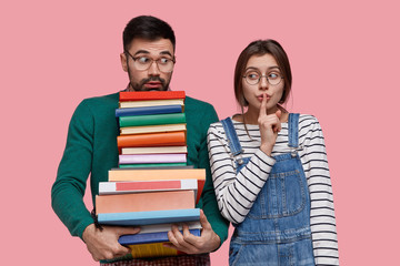 Shh, dont tell our secret to anybody. Beautiful dark haired woman makes silence gesture, dressed in striped jumper and overalls, stands closely to her groupmate who holds many books, prepare for test