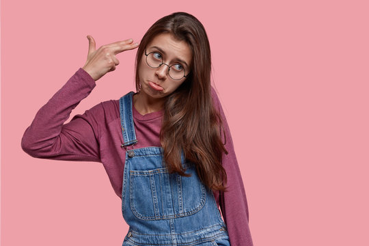 Upset dark haired woman shoots and kills herself, points with fingers at temple, purses lower lip in displeasure, wears transparent glasses and overalls, isolated over pink background. Suicide gesture