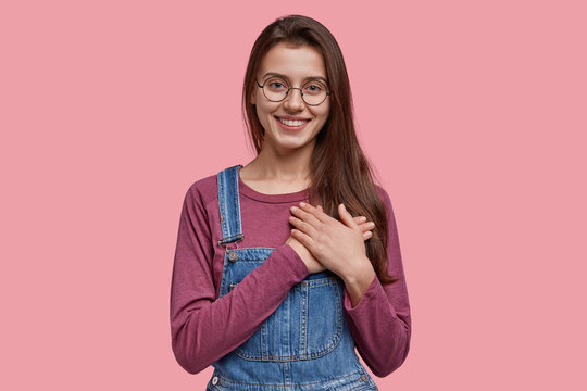 Kind European woman with pleasant smile, express favour, keeps both hands on chest, being kind hearted and honest, dressed in denim overalls, round optical glasses, isolated over pink background