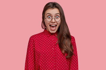 Attractive brunette overjoyed woman opens mouth and yells from joy, wears pink polka dot blouse,...