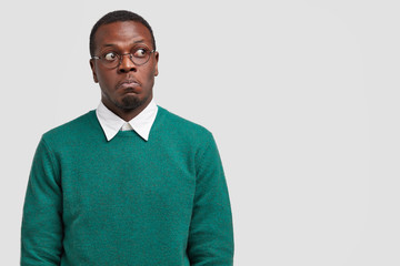 Puzzled dark skinned man looks thoughtfully and with bewilderment, focused aside, dressed in casual sweater, optical glasses, models over white studio wall with free space for your text or information