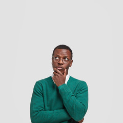 Vertical shot of contemplative thoughtful dark skinned man keeps hand under chin, looks thoughtfully upwards, dressed in green sweater, poses over white background with free space for your text