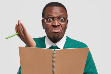 Confused black young man has hesitant expression, doesnt know waht to write, holds textbook and pen, prepares story about his holidays, wears optical glasses, isolated over white background.