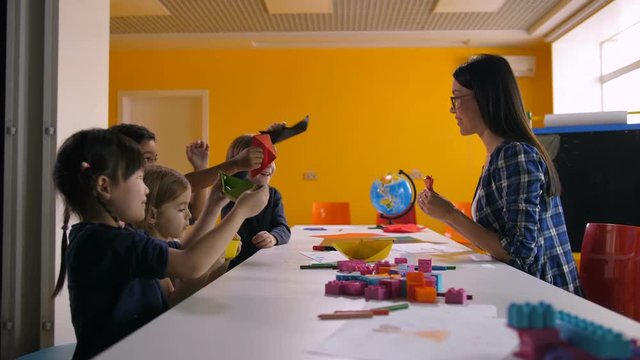 Joyful carefree multi ethnic diverse children playing with handmade origami toys at craft lesson. Happy kindergarten teacher and little students having fun in preschool class.