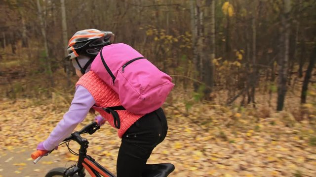 One caucasian children rides bike road in autumn park. Little girl riding black orange cycle in forest. Kid goes do bicycle sports. Biker motion ride with backpack and helmet. Mountain bike hardtail.