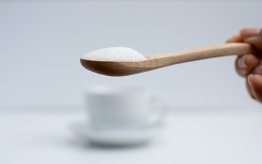 Hand holding brown wooden sugar spoon in front of blurred white coffee cup in the background