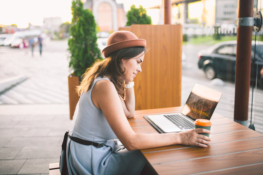 Beautiful young caucasian woman sitting on a terrace in a cafe in summer at a wooden table in a hat and a plate uses technology, working behind a laptop booking hotels trip in the summer