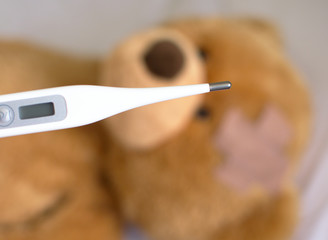 White digital thermometer in the blurred sick brown teddy bear  background 